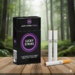 บุหรี่ Lucky Strike Purple ซองสีดำวงกลมม่วง พร้อมมวนบุหรี่เม็ดบีบวางบนโต๊ะไม้ฉากหลังป่าสีเขียว