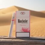 ซองบุหรี่ Manchester Pink SuperSlim สีขาวชมพู วางบนขอนไม้กลางทะเลทราย แสงแดดสีทอง ดีไซน์ทันสมัย