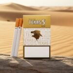 ซองบุหรี่ Texas 5 Gold วางบนขอนไม้ในทะเลทราย แสงสีทองพรีเมียม แสดงดีไซน์ซองสีทองและมวนบุหรี่